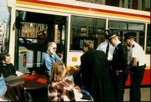 Dr Bill Albert handcuffed to a kneeling bus accompanied by colleagues and attended by police officers with bolt cutters and a man from the Ministry in September 1996